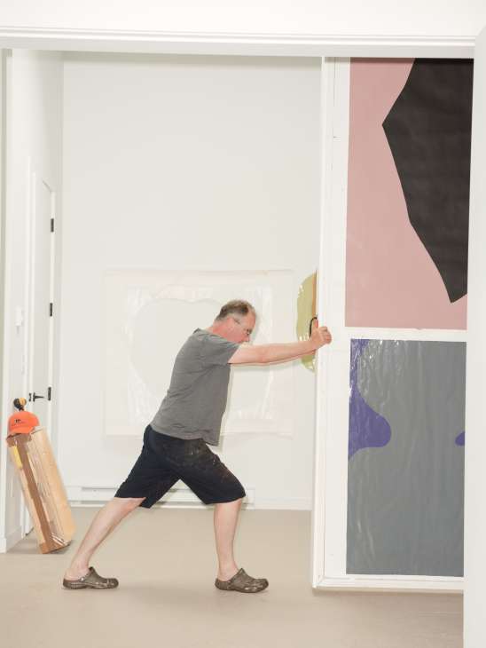 Artist Gary Hume adjusting artwork in his studio, surrounded by colorful paintings with abstract shapes in pink, black, purple and grey.