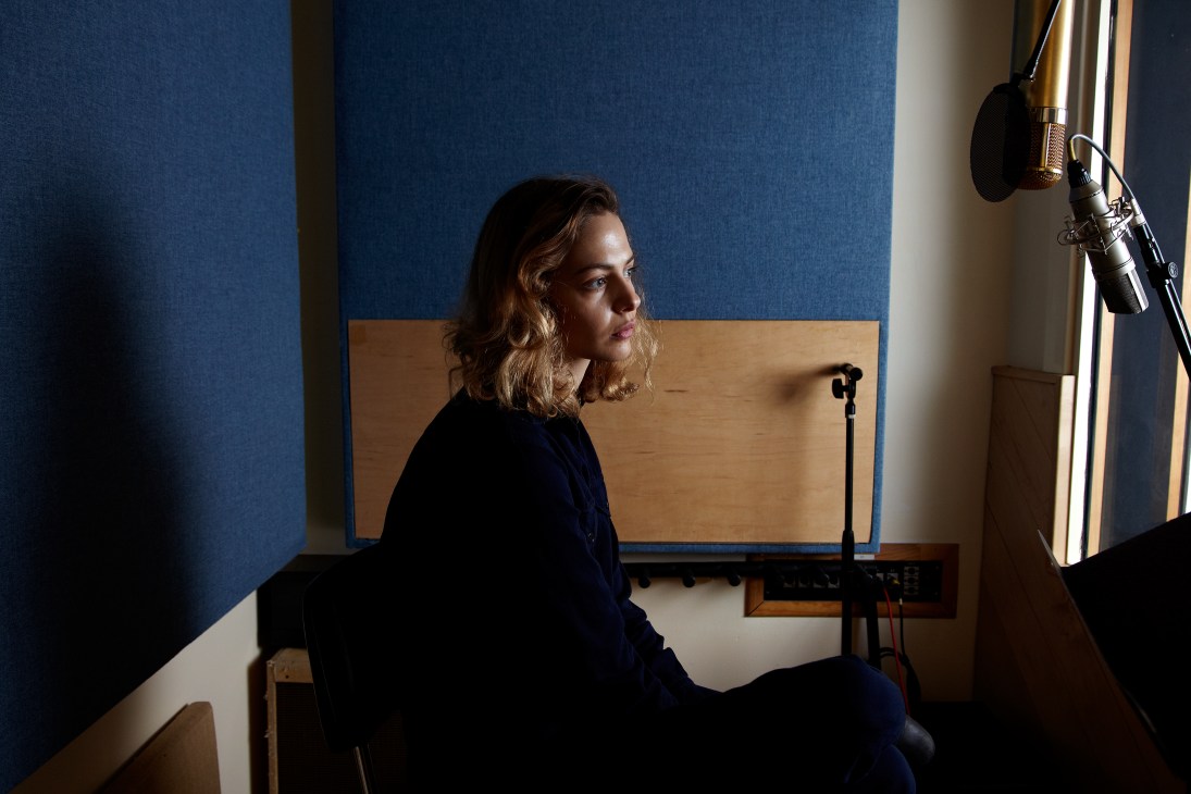 Hannah Cohen sits in a Manhattan studio with blue and tan walls, positioned near a microphone and professional recording equipment.