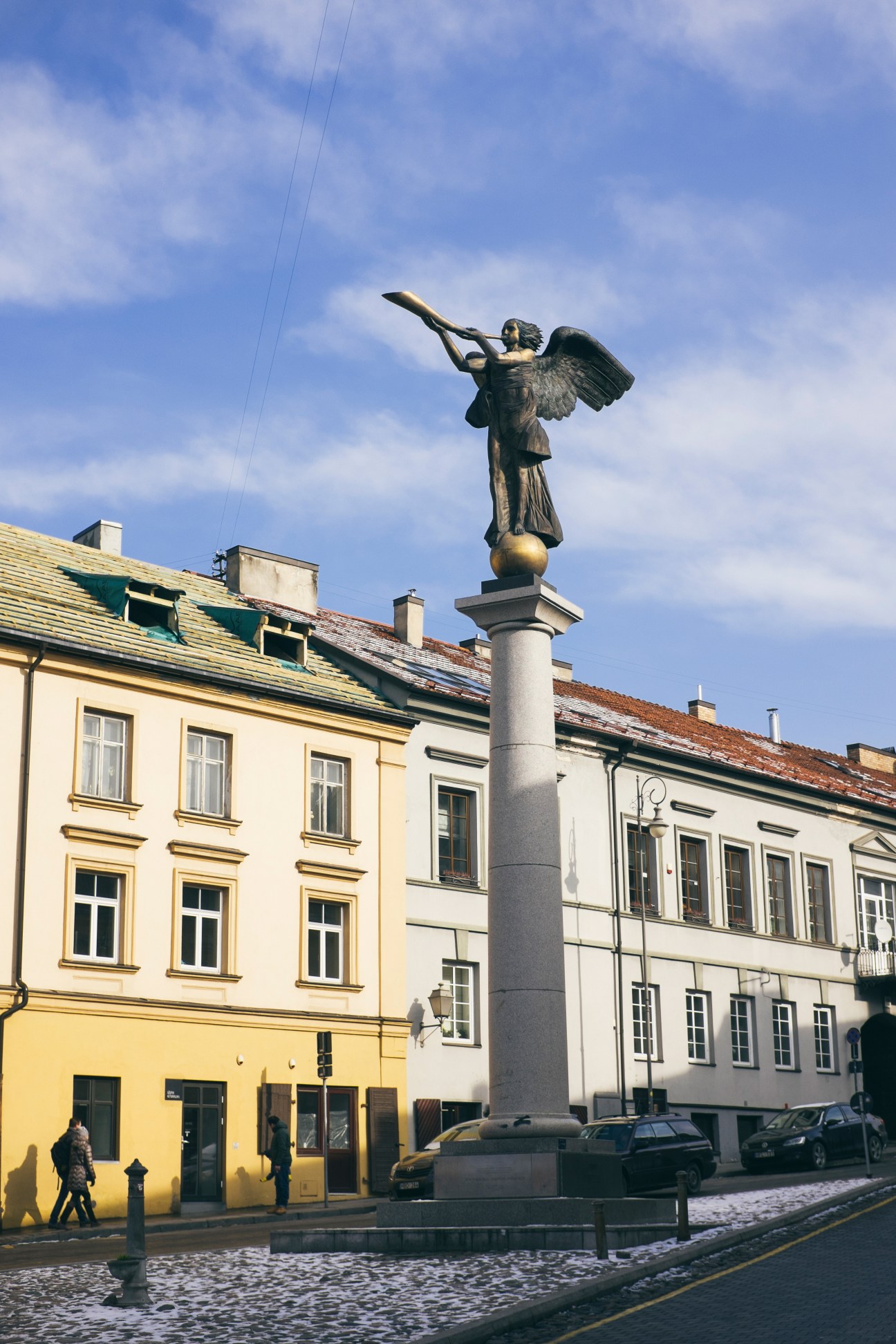 Bronze angel statue on a pedestal in Uzupis district with colorful historic buildings behind it.