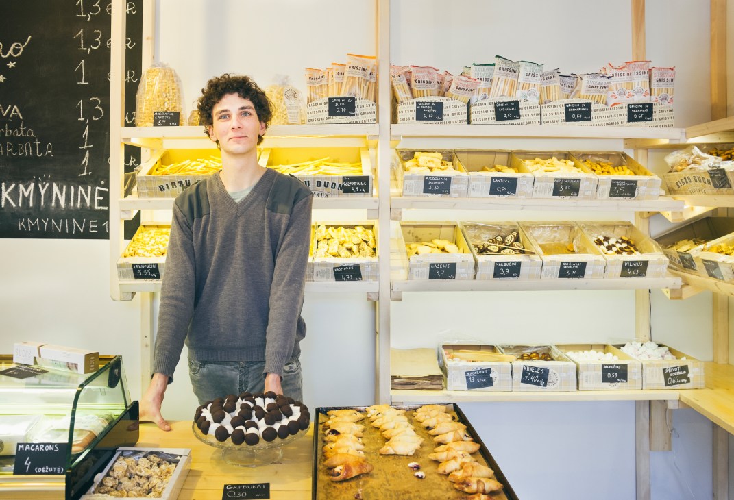 Vytenis Daubaras stands behind the counter at Kmynine bakery surrounded by pastries and baked goods on shelves.