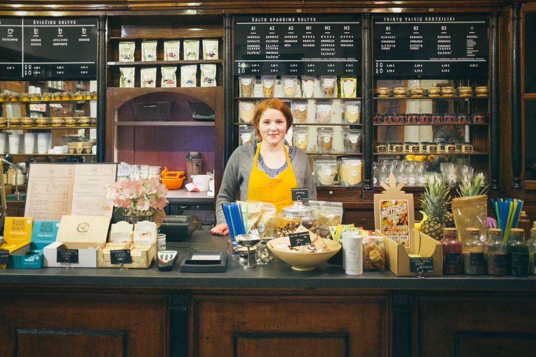 Woman in yellow apron stands behind counter at Vaisine shop with shelves of goods.