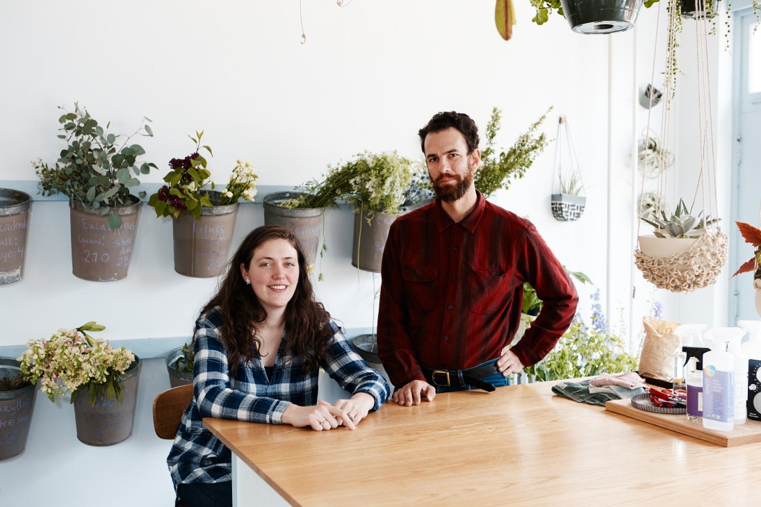 Mairead and Seth Travins, owners of Flowerkraut, stand behind a wooden counter in their bright studio space filled with potted plants and hanging succulents.