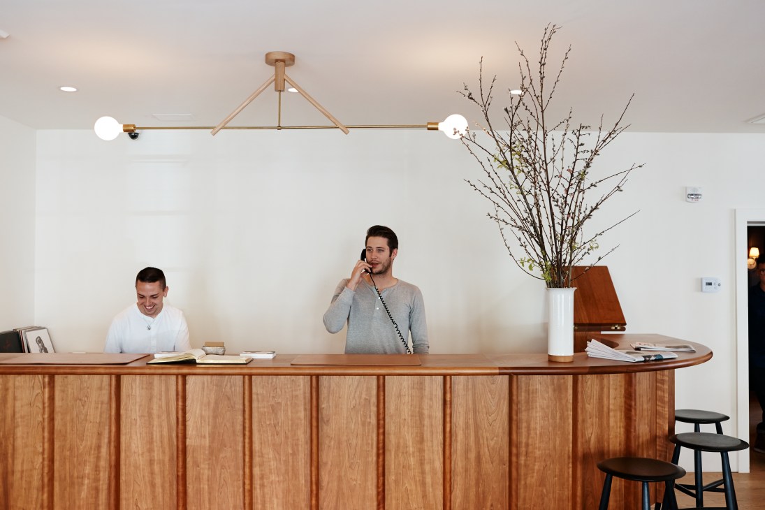 Two staff members at the reception desk of Rivertown Lodge, a wooden counter with modern brass lighting fixture above.
