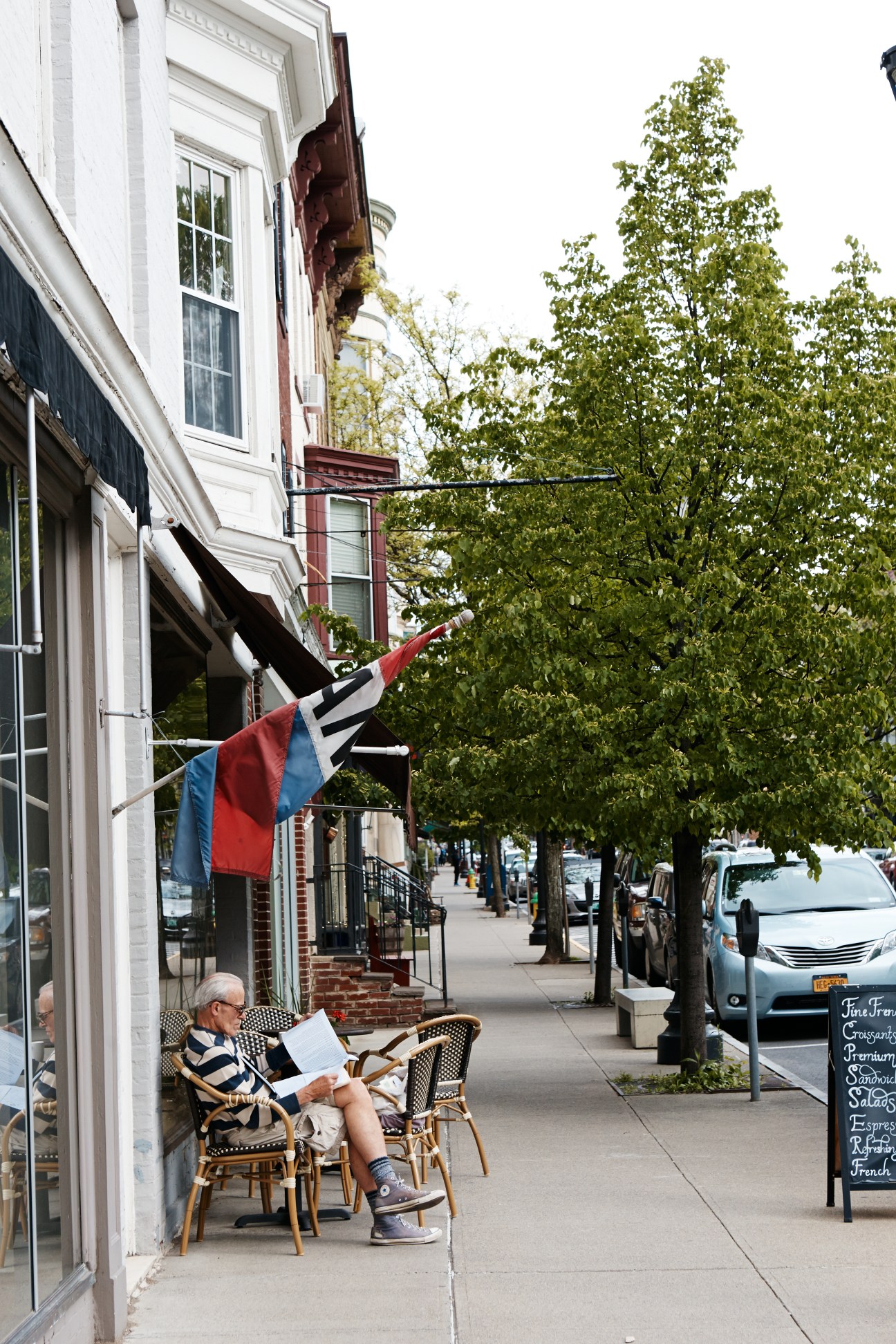 Man reading newspaper seated outside a storefront on Warren Street lined with trees and storefronts.