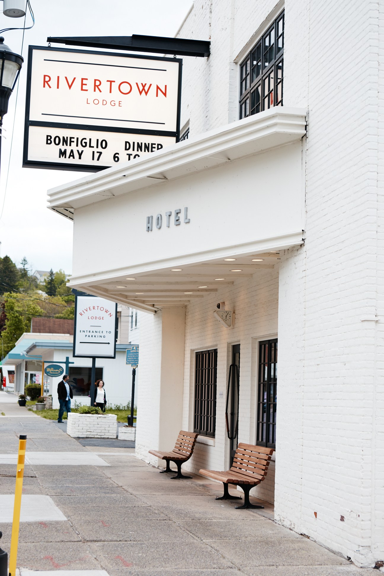 White-painted hotel building with red Rivertown Lodge signage and wooden benches outside entrance.