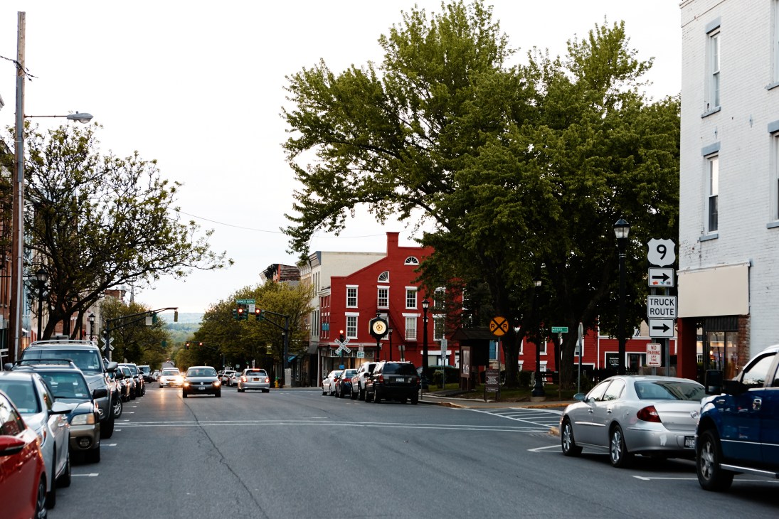 Warren Street in Hudson lined with parked cars, a red brick building, and leafy trees under a cloudy sky.