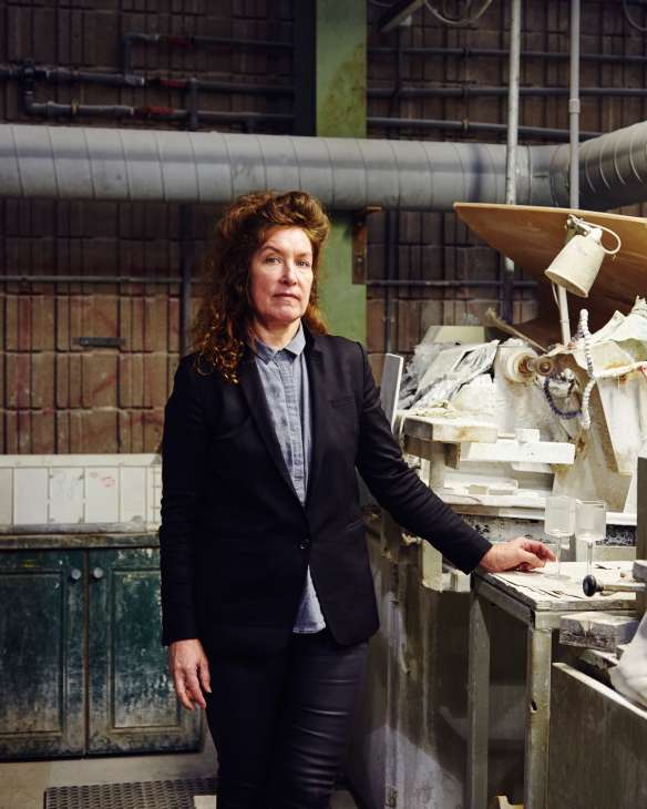 Anike Tyrrell stands in an industrial workspace with glassmaking equipment and finished glass pieces behind her.