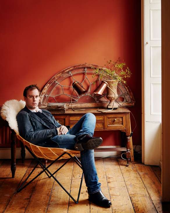 Man sitting in a chair against a red wall with wooden furniture and copper vessels displayed behind him.