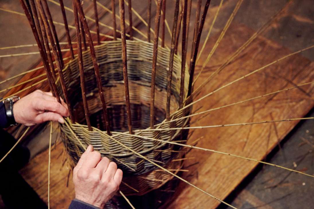Hands weaving willow strips in a traditional basket-making craft technique.