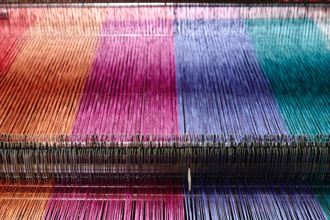 Colorful linen swatches on a loom showing vibrant gradients from orange through pink, purple, blue to teal with black threads.