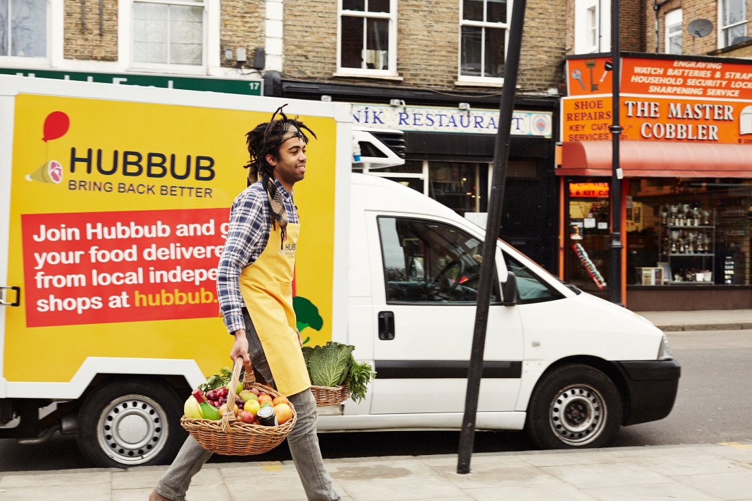 Man in yellow apron holding basket of fresh produce in front of Hubbub delivery vehicle.