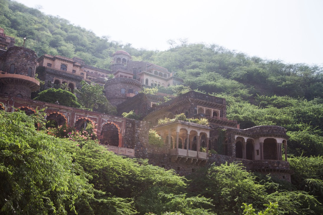 Neemrana Fort Palace Hotel, a historic multi-story palace structure with terracotta and stone architecture nestled among lush green hillside vegetation.