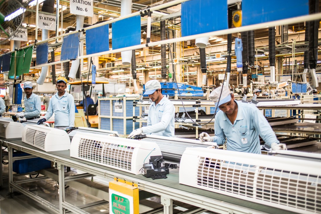 Workers in light blue uniforms and caps assemble air conditioning units on a conveyor line at the Daikin factory in Neemrana.