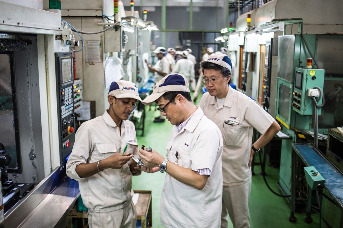 Workers in white uniforms and Nissin Brake caps inspect components on a factory floor with green flooring.