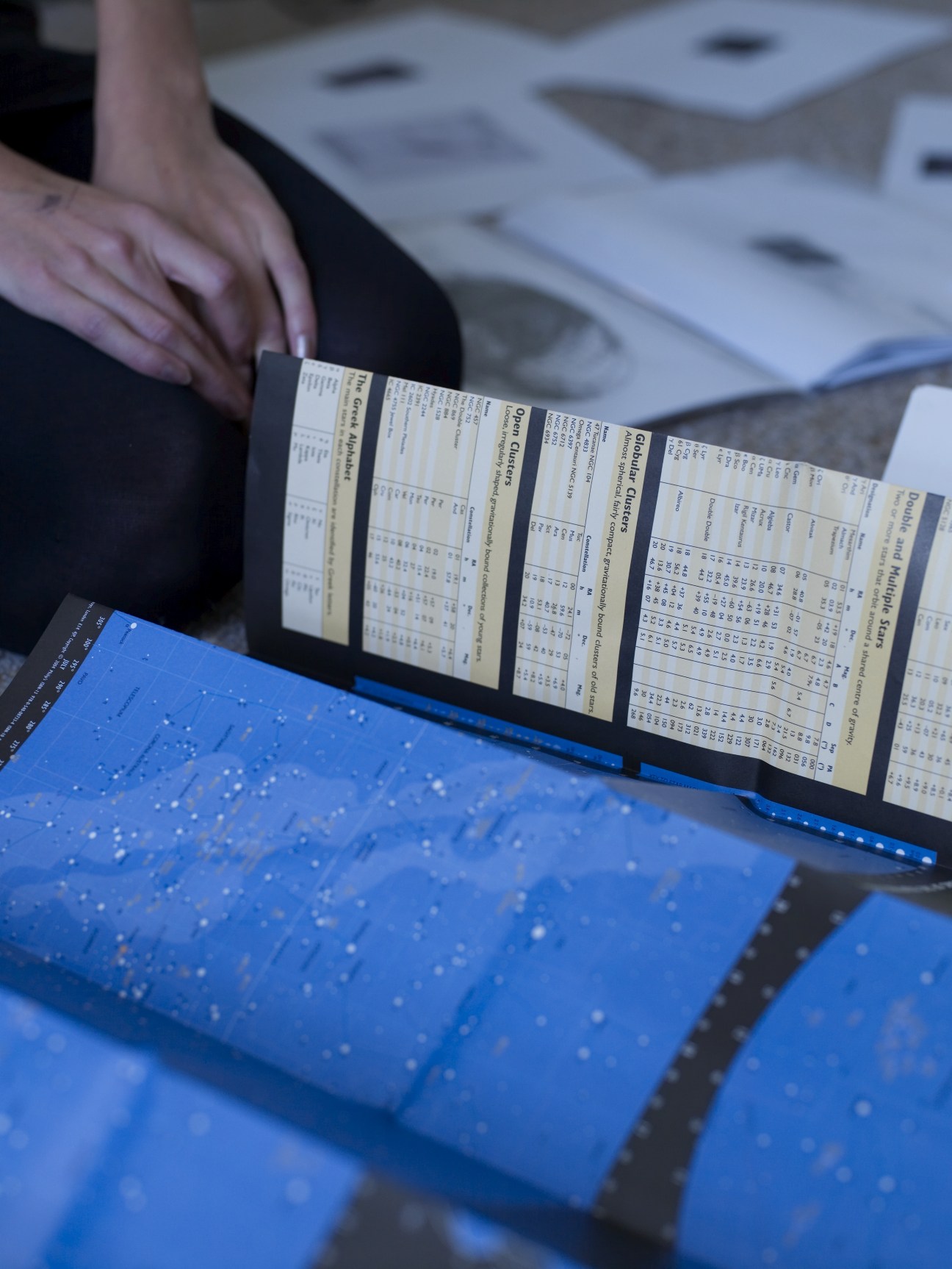 Katie Paterson holds open a blue star map with her hand while reviewing astronomical charts at her desk.