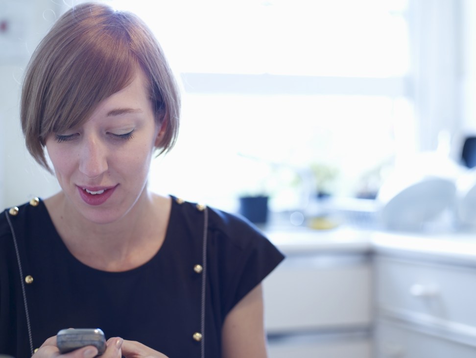 Katie Paterson holds a mobile phone in her hands while looking down at it in her bright home office.