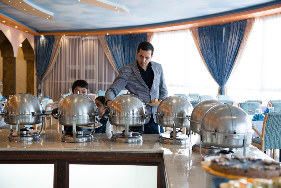 Hotel staff member serving buffet lunch under metallic domes in an elegant dining room with blue curtains.