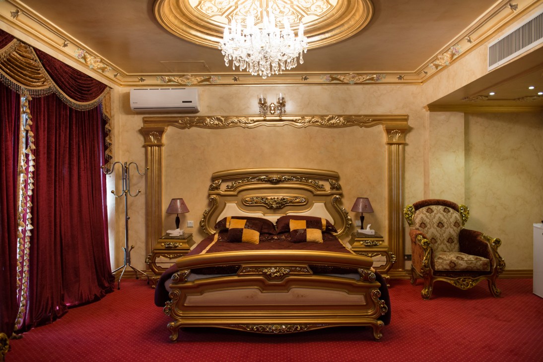 Ornate gold-trimmed bedroom with elaborate carved bed, burgundy upholstery, crystal chandelier and red carpet.