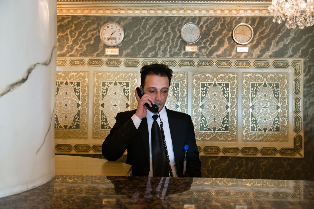 Man in business suit speaking on telephone at hotel reception desk with decorative patterned wall and clocks in background.