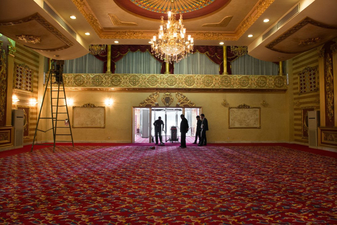 Hotel staff cleaning a large ornate ballroom with gold-trimmed walls, red patterned carpet, and a crystal chandelier.