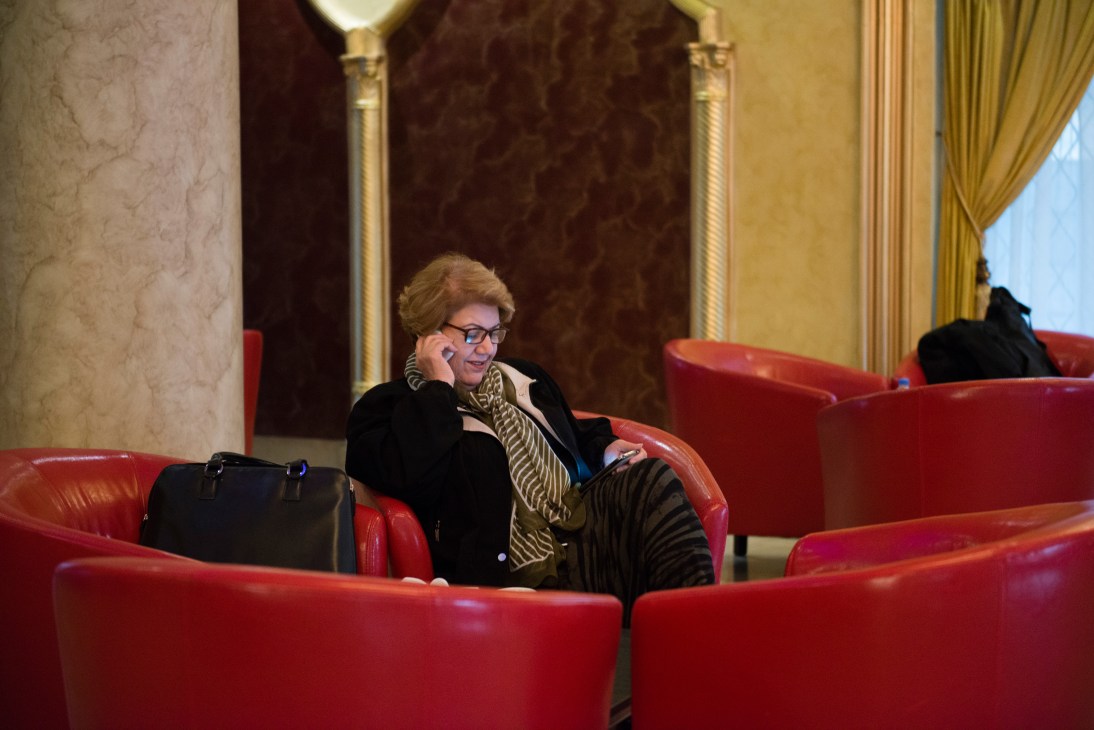 Woman in glasses and black jacket sits in red leather chair in ornate hotel lobby, talking on phone with briefcase beside her.