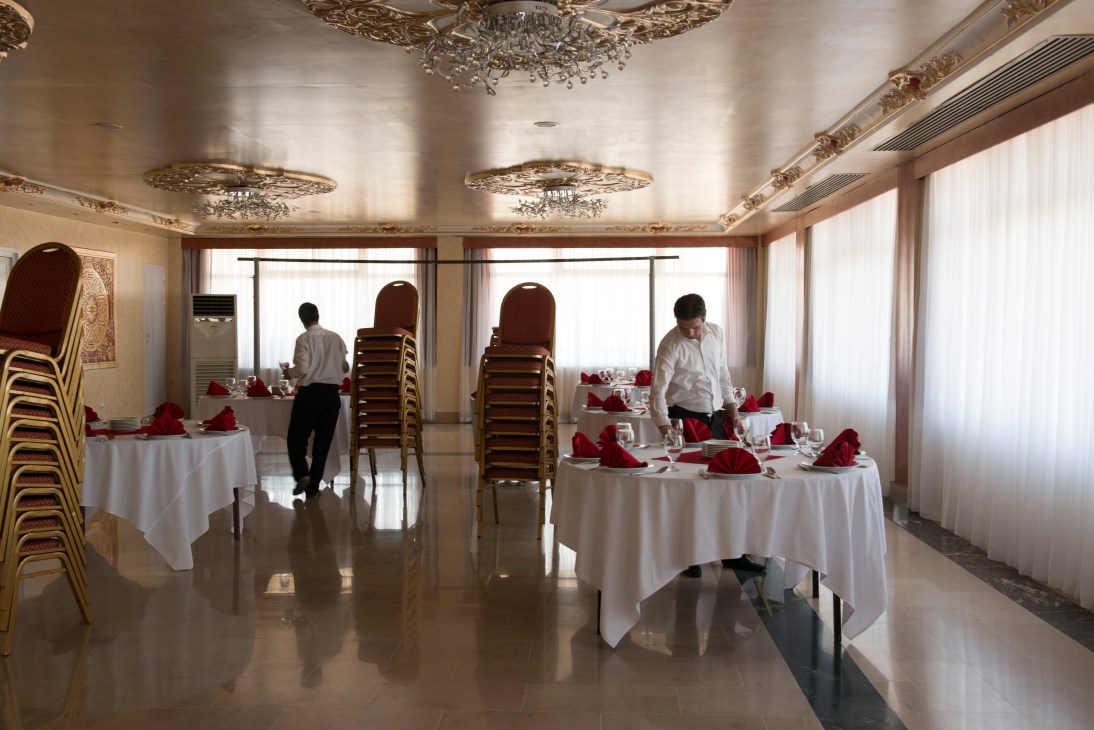 Hotel staff setting up round tables with white linens and red napkins in an ornate ballroom.