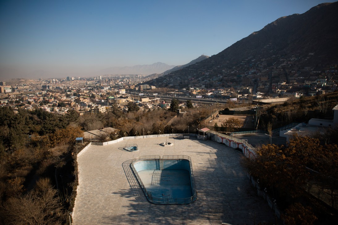 Empty swimming pool on a hotel terrace overlooking Kabul's sprawling cityscape and snow-capped mountains.