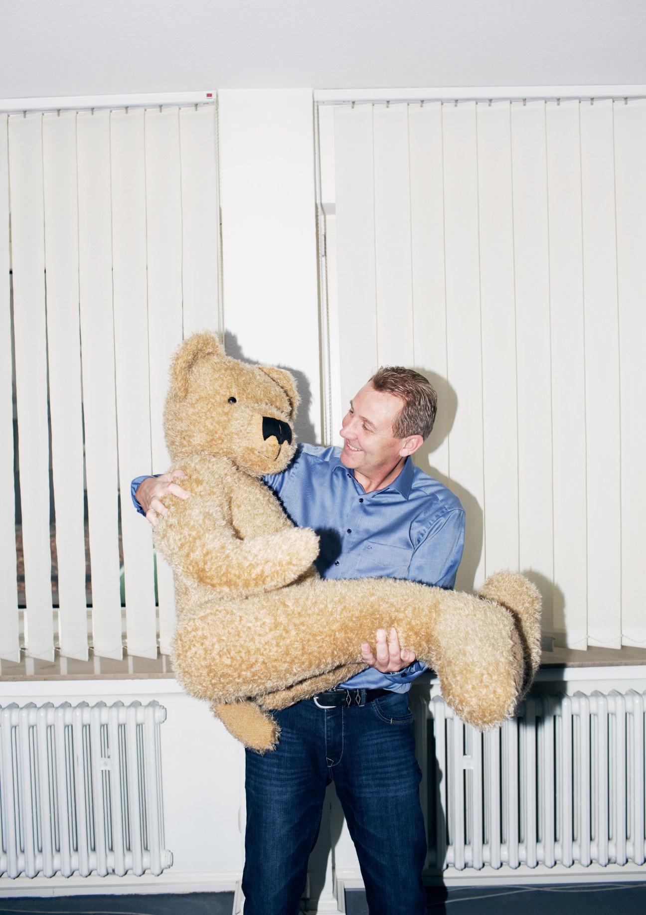 Bernhard Wanning, managing director of Steiff Schulte, holds a large tan teddy bear in an office with vertical blinds.