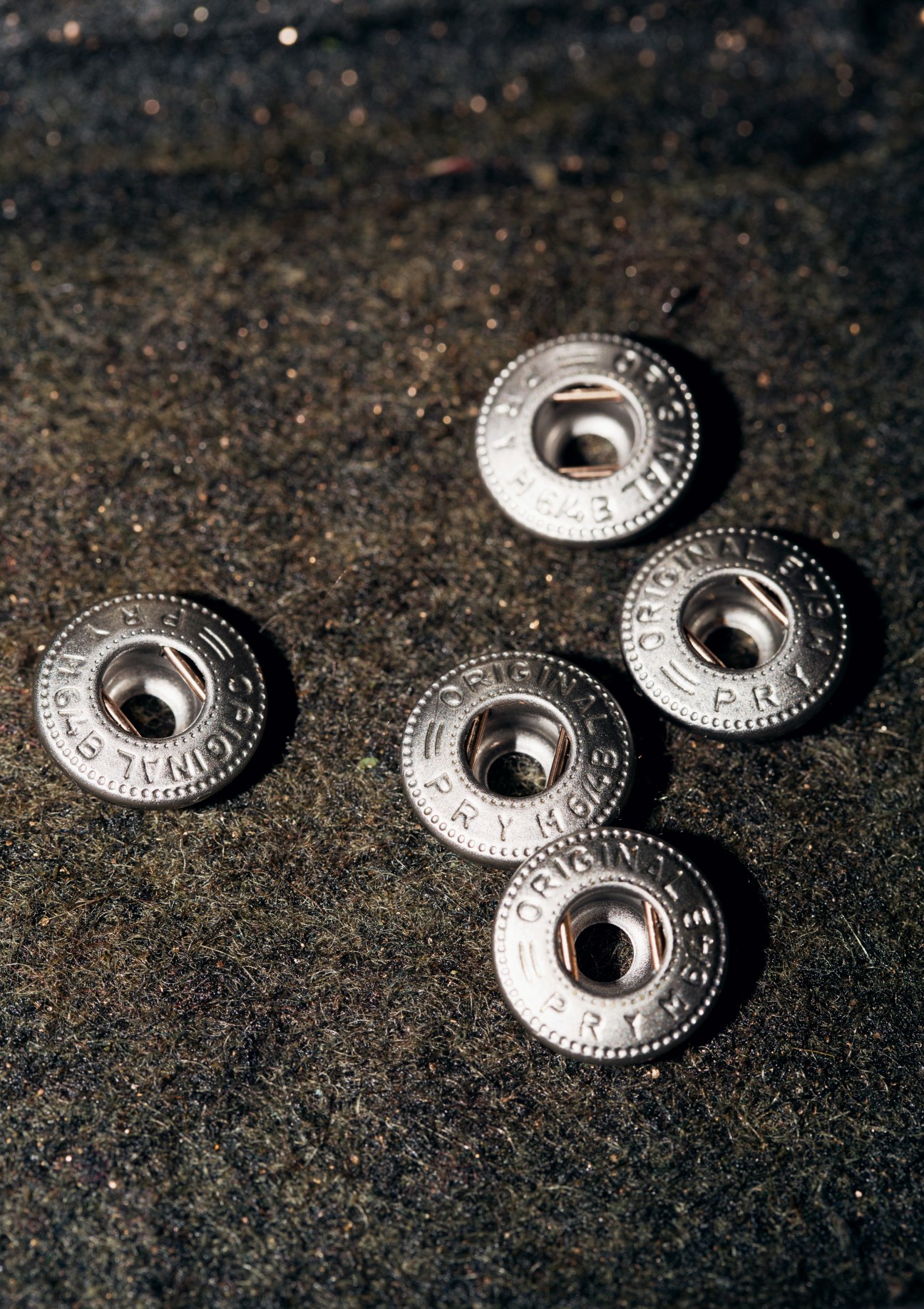 Five Prym snap fasteners arranged on a textured dark surface, showing the finished metal poppers before assembly.