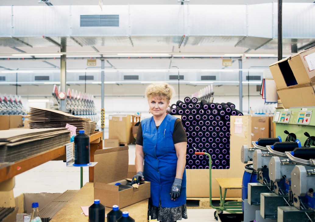 Woman in blue vest standing in textile factory with yarn bobbins and production equipment visible behind her.