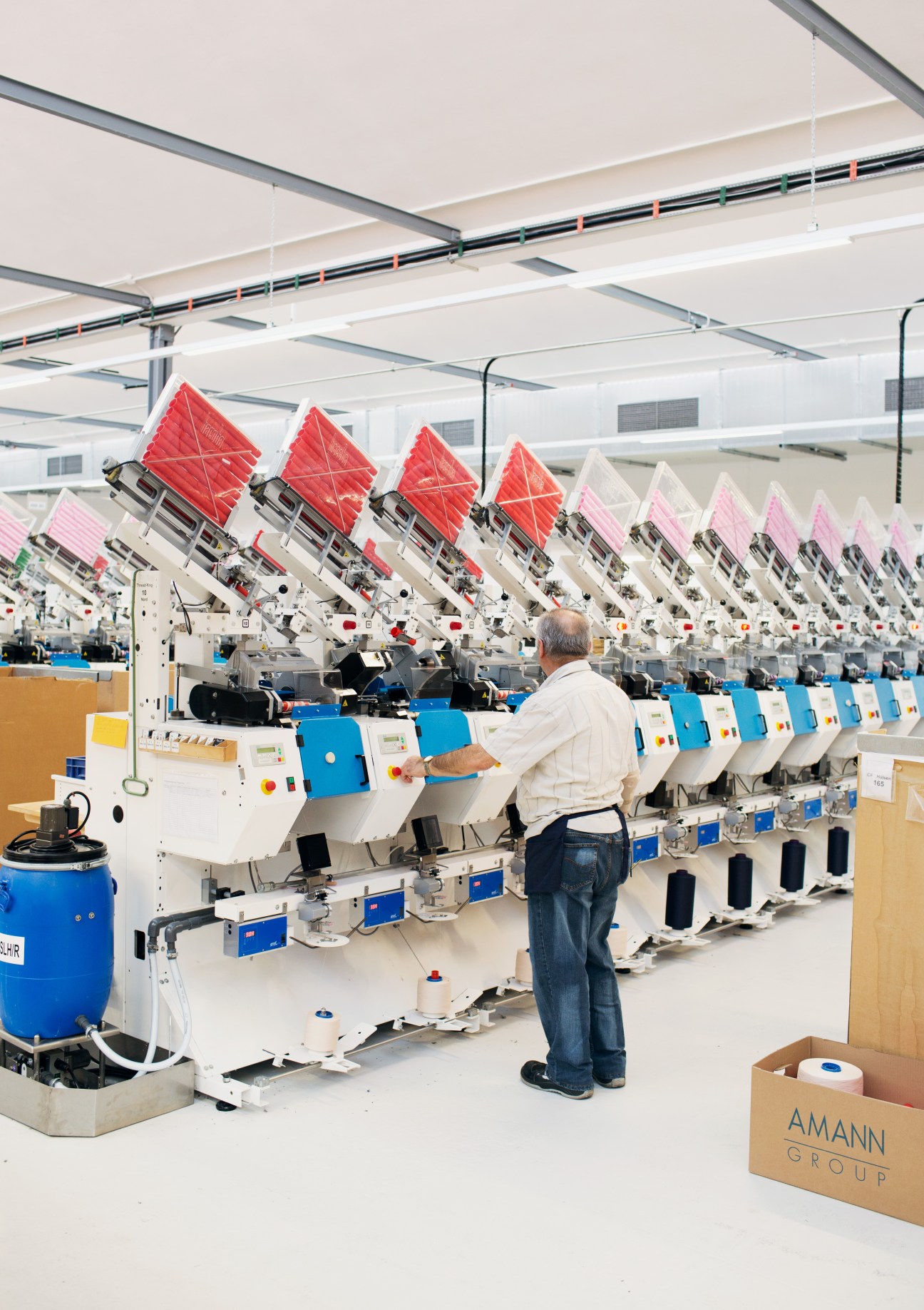 Worker operating yarn winding machines at Amann & Söhne's Augsburg factory with red and pink bobbins.