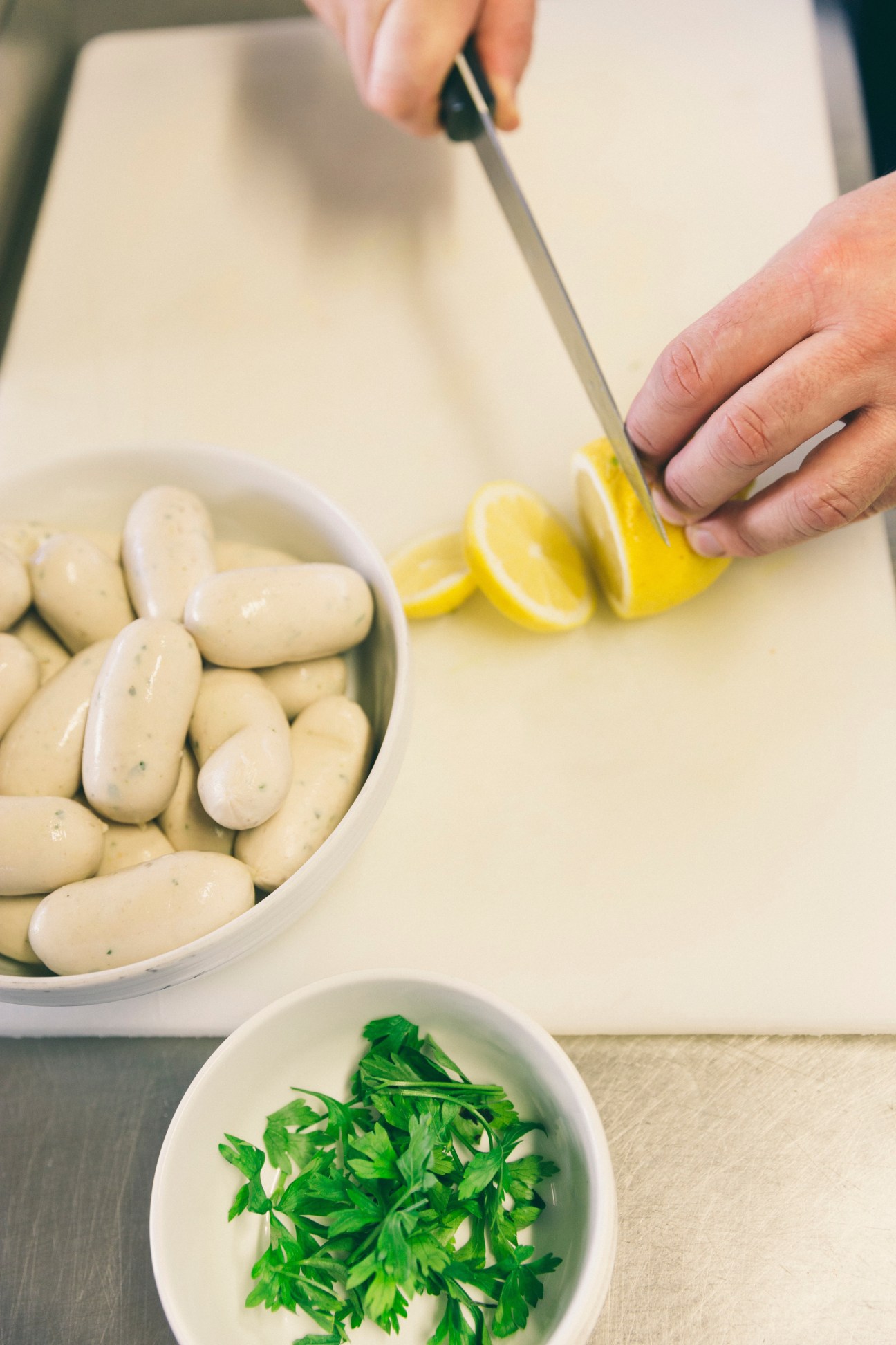 Hands slicing a lemon over a bowl of Weisswurst sausages with parsley in a small dish nearby.