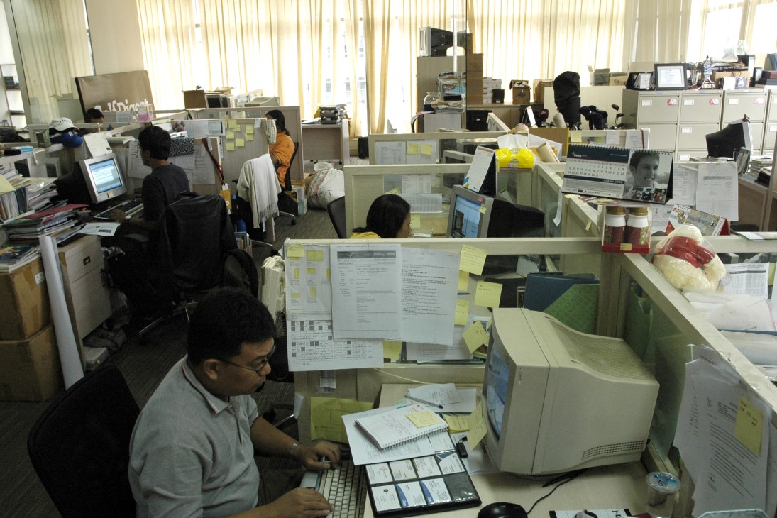 Journalists working at desks in a busy newsroom office with computers and office cubicles.