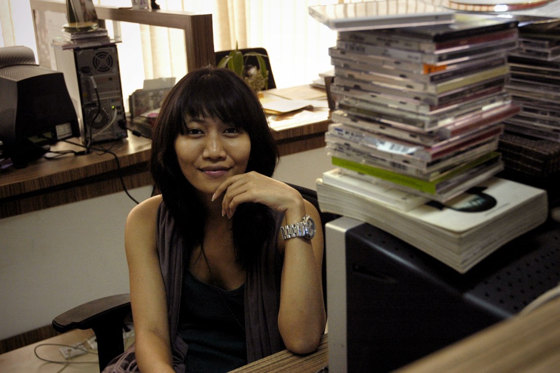 Putri Avianti, beauty editor at Mahaka's A+ magazine, sits at her desk surrounded by stacked magazines and media materials.