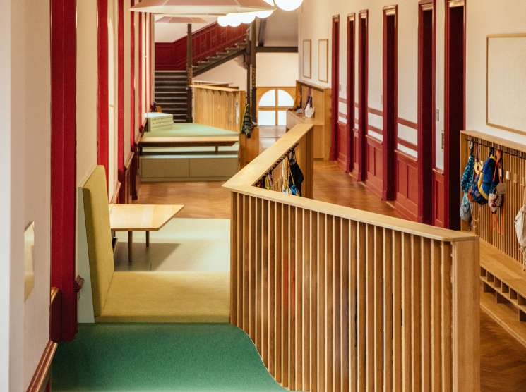 Modern school corridor with green carpeted flooring, red doors, wooden railings, and pendant lights overhead.