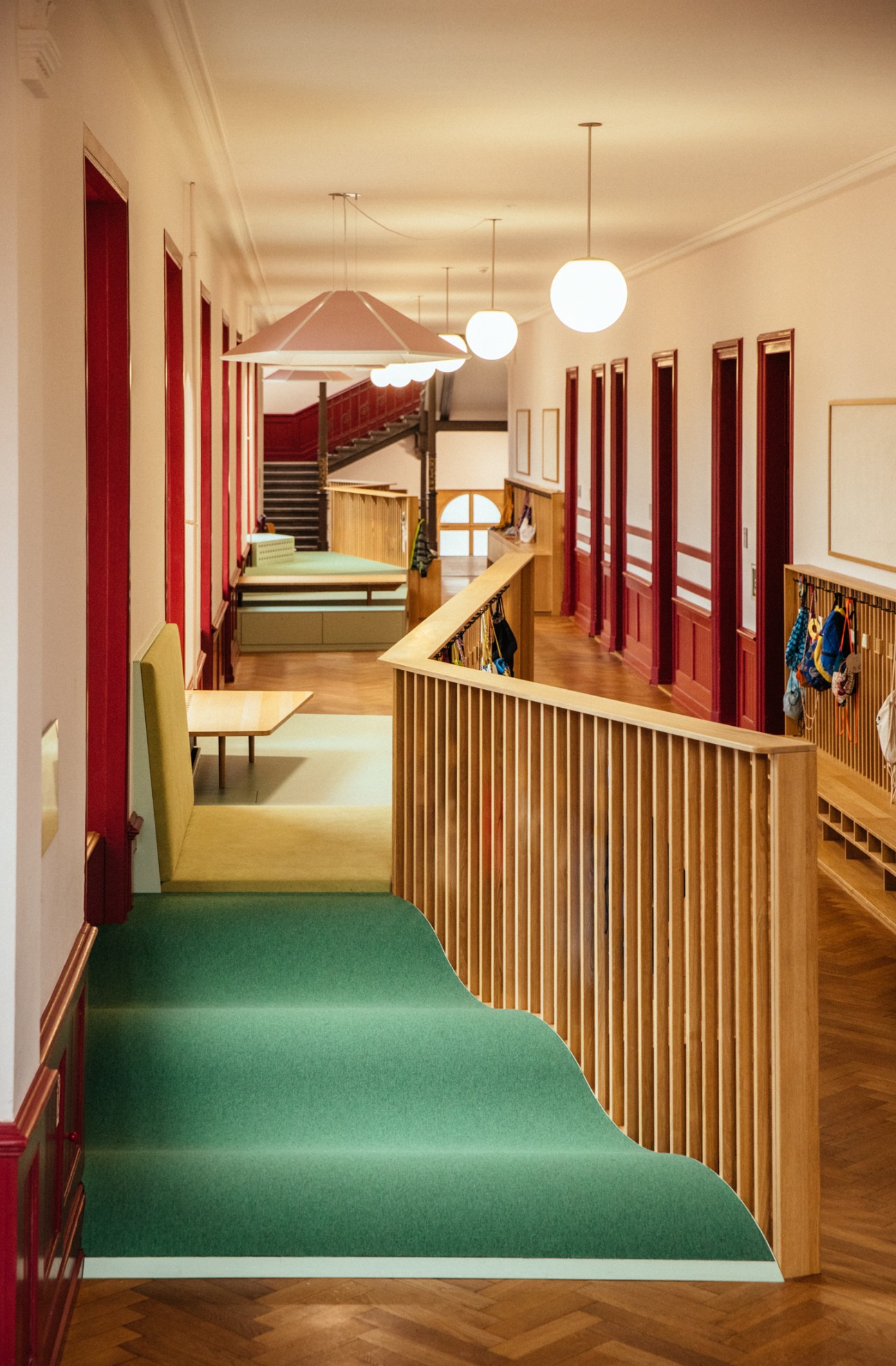 Modern school corridor with green carpeted flooring, red doors, wooden railings, and pendant lights overhead.