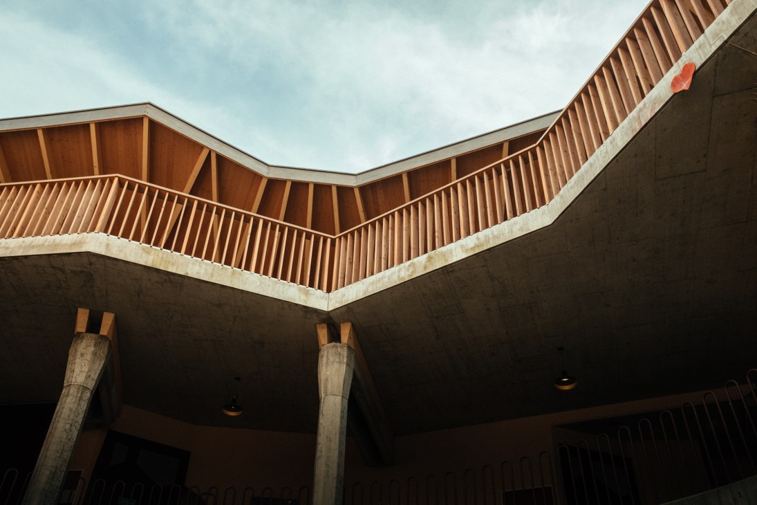 Upward view of angular larch-wood ceiling with corrugated metal panels and concrete pillars in Geneva school.