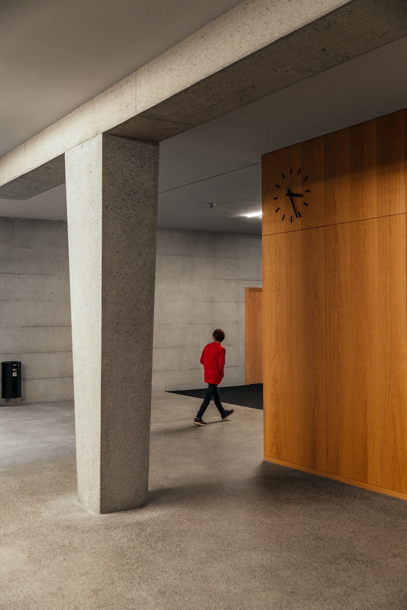Child in red jacket walking through Zofingen primary school entrance hall with concrete pillars and wooden wall featuring clock.