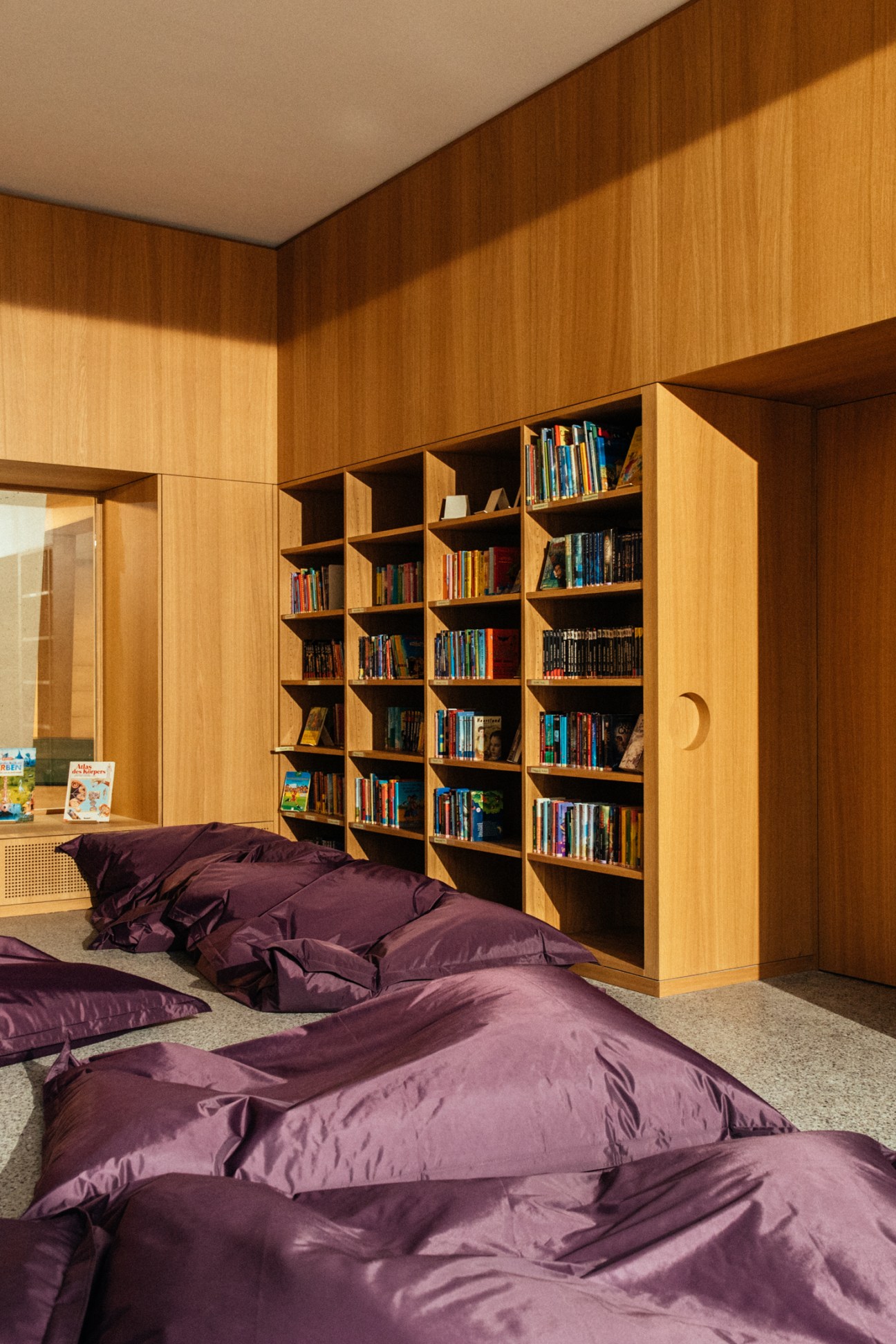 Wooden library shelving with books displayed against warm oak-panelled walls beside purple floor cushions.