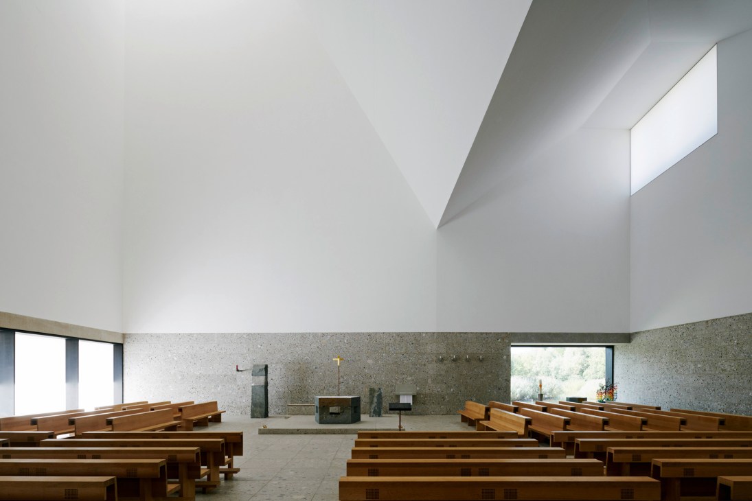 Interior of Church of the Blessed Father Rupert Mayer with wooden pews, stone altar, and skylight casting natural light on white walls.