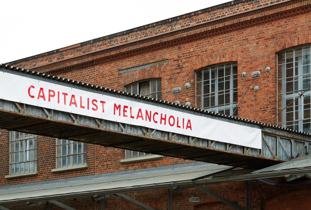 Red brick industrial building with 'Capitalist Melancholia' exhibition banner in Hall 14 at Leipzig's Spinnerei complex.
