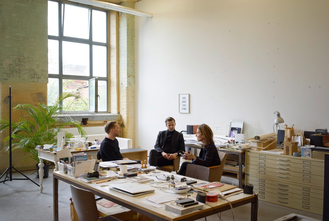Two people in conversation in a bright, minimalist studio space with large windows and work tables.