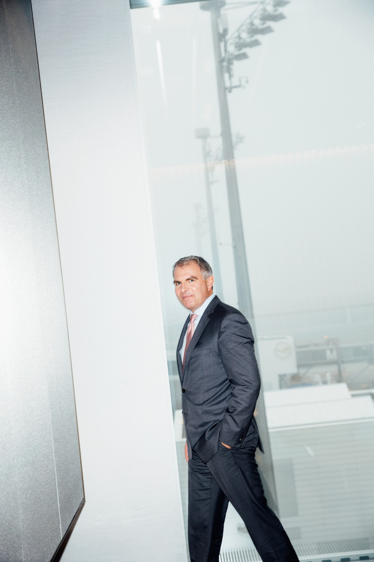 Lufthansa CEO Carsten Spohr in dark suit and pink tie standing in modern white First Class lounge at Munich Airport.
