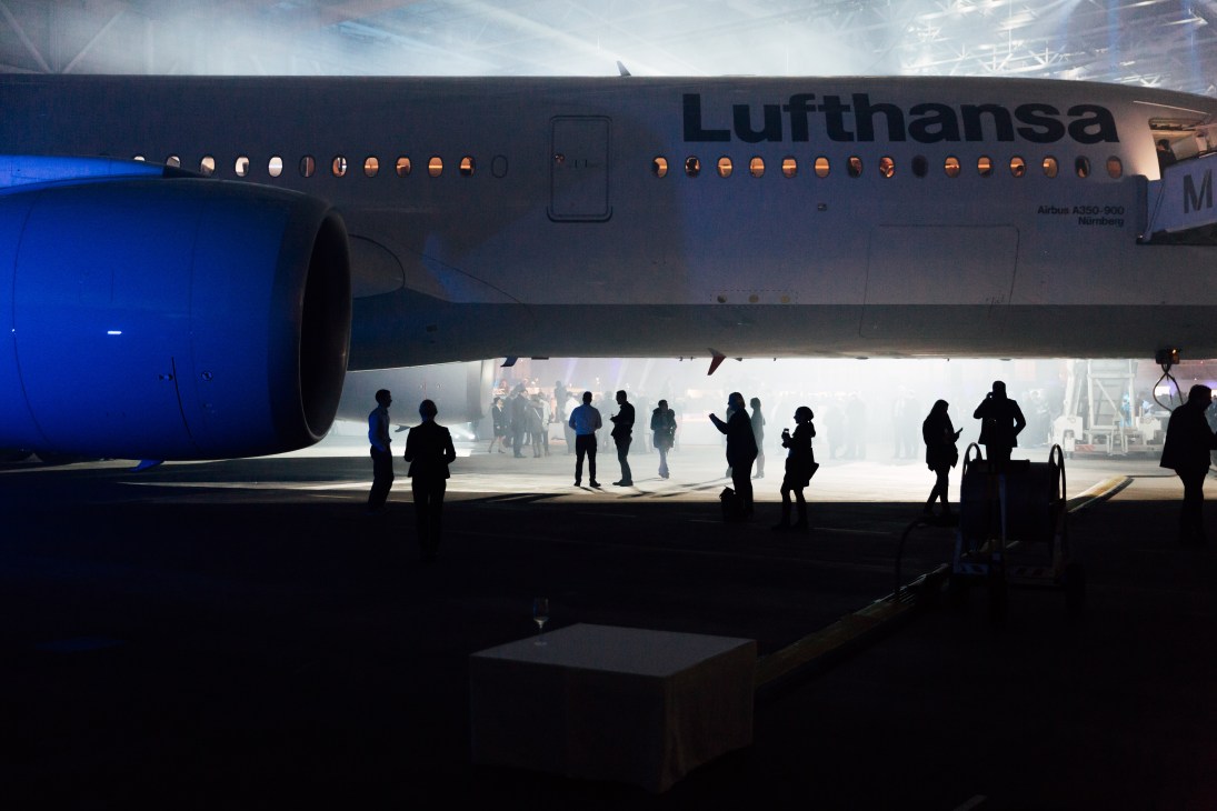 Guests and staff silhouetted beneath a Lufthansa Airbus A350-900 aircraft in a hangar during an unveiling event.