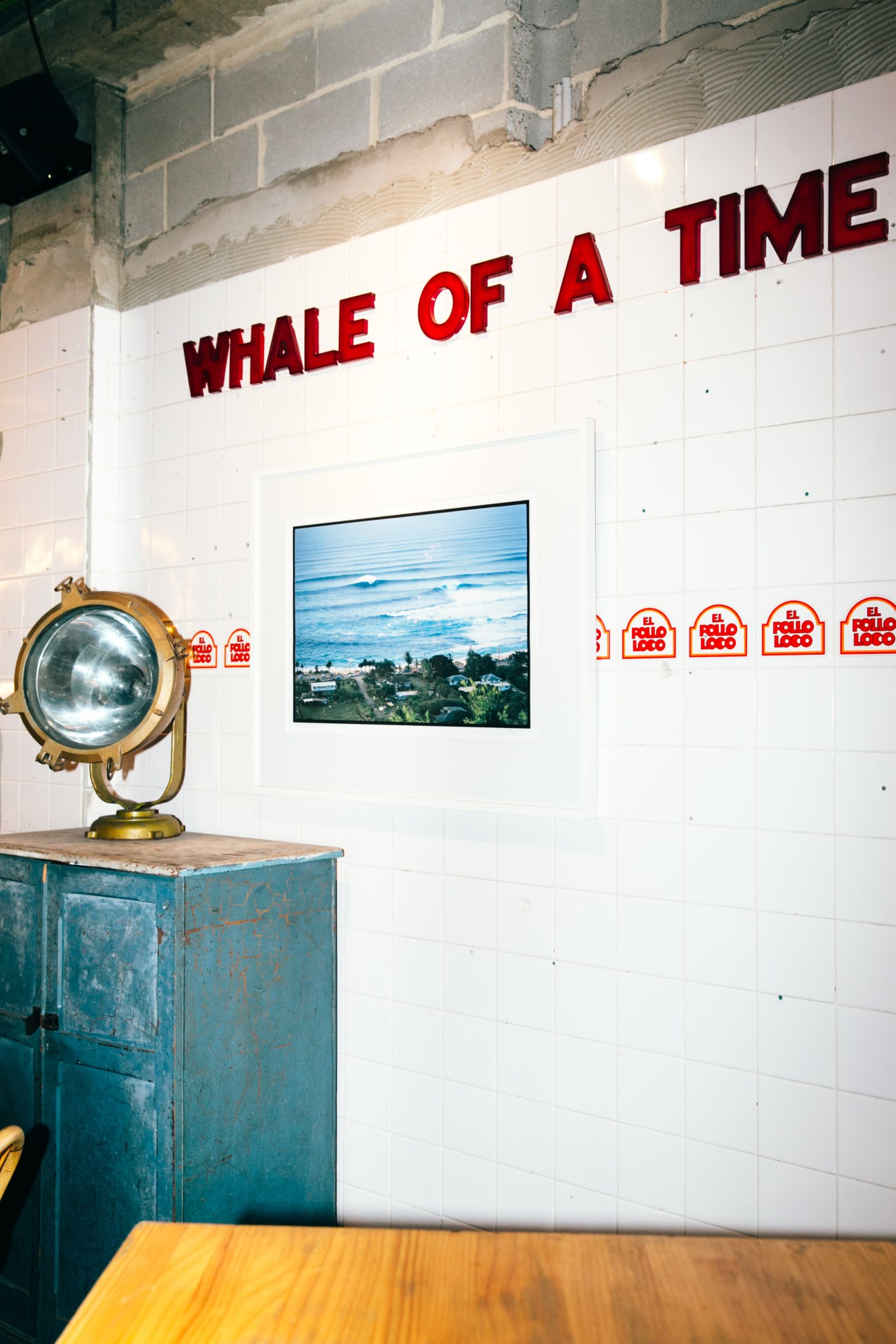 Nautical interior of Coogee Pavilion with brass spotlight, white tile wall, and red "Whale of a Time" signage above a coastal landscape photograph.
