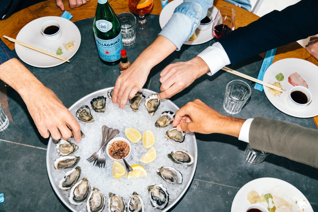 Multiple hands reaching for oysters on ice at a restaurant table with San Pellegrino and glasses of wine.