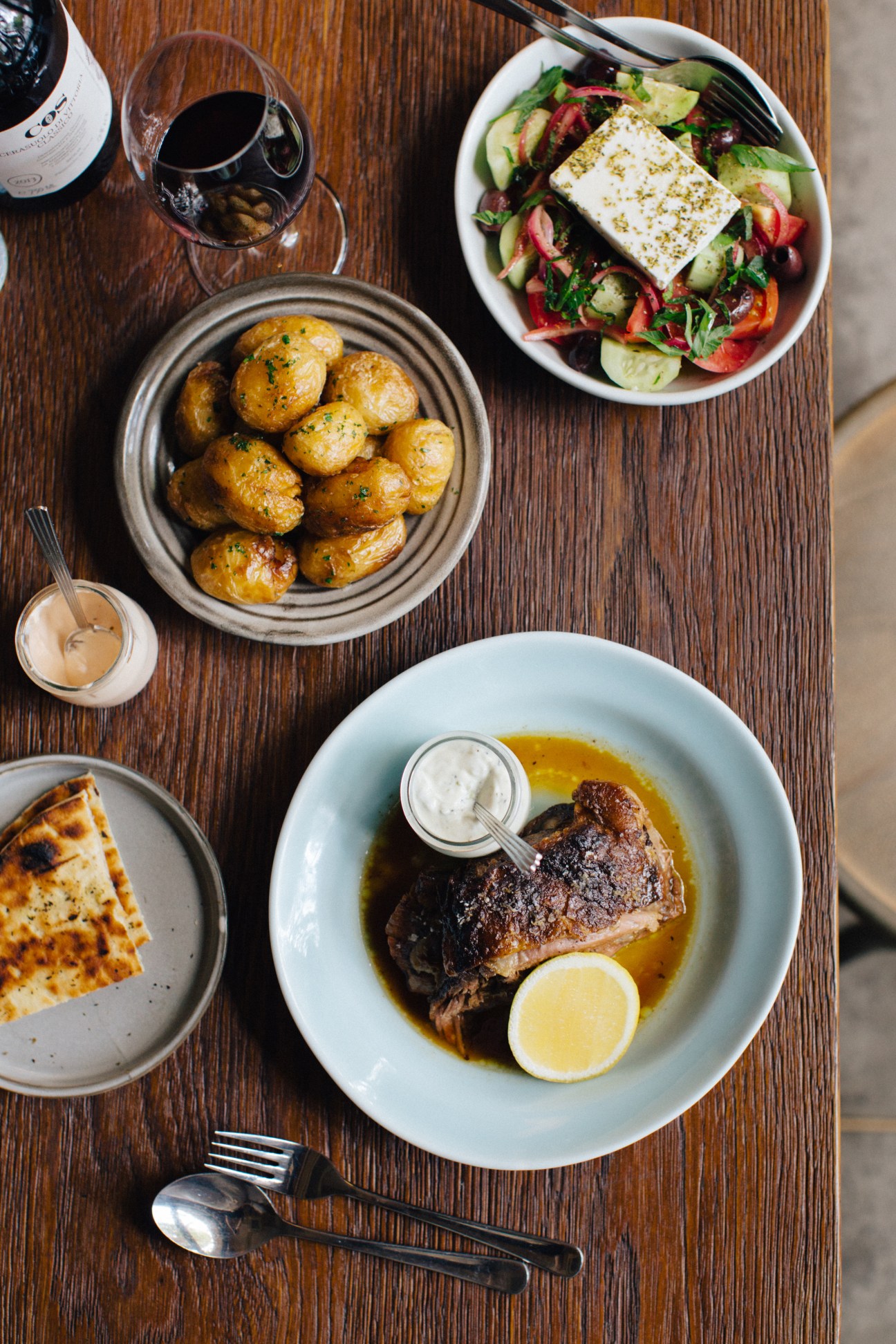 Overhead view of Greek dishes at The Apollo including roasted potatoes, salad with feta, lamb, and pitta bread on a wooden table.