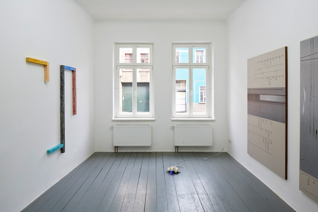 Bright gallery space with large windows, painted artworks on walls, and grey wooden flooring.