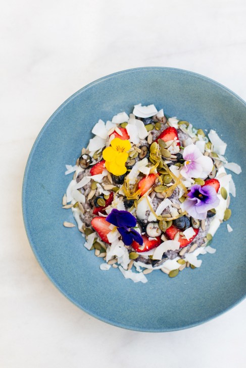 Chia black rice pudding garnished with edible flowers, coconut, and pumpkin seeds on a blue plate.