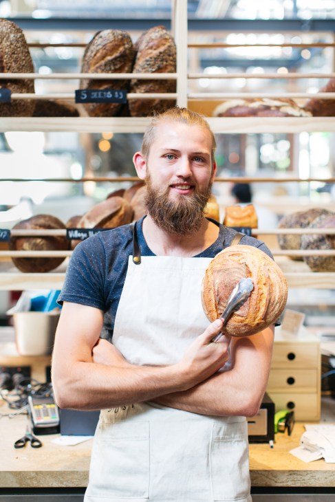 Baker holding a round loaf of bread in front of shelves displaying various artisan breads at Dust bakery.